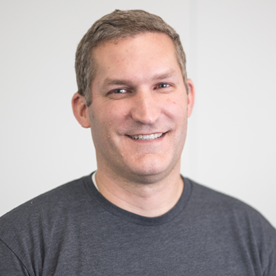 team-greg Smiling man in gray t-shirt against white background, professional headshot.