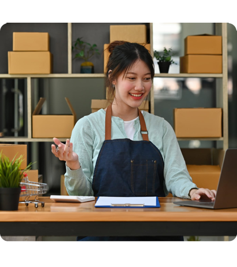 Smiling woman in apron at desk with laptop and cardboard boxes behind her.