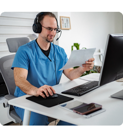 Healthcare professional in blue scrubs using tablet at desk with laptop and headset